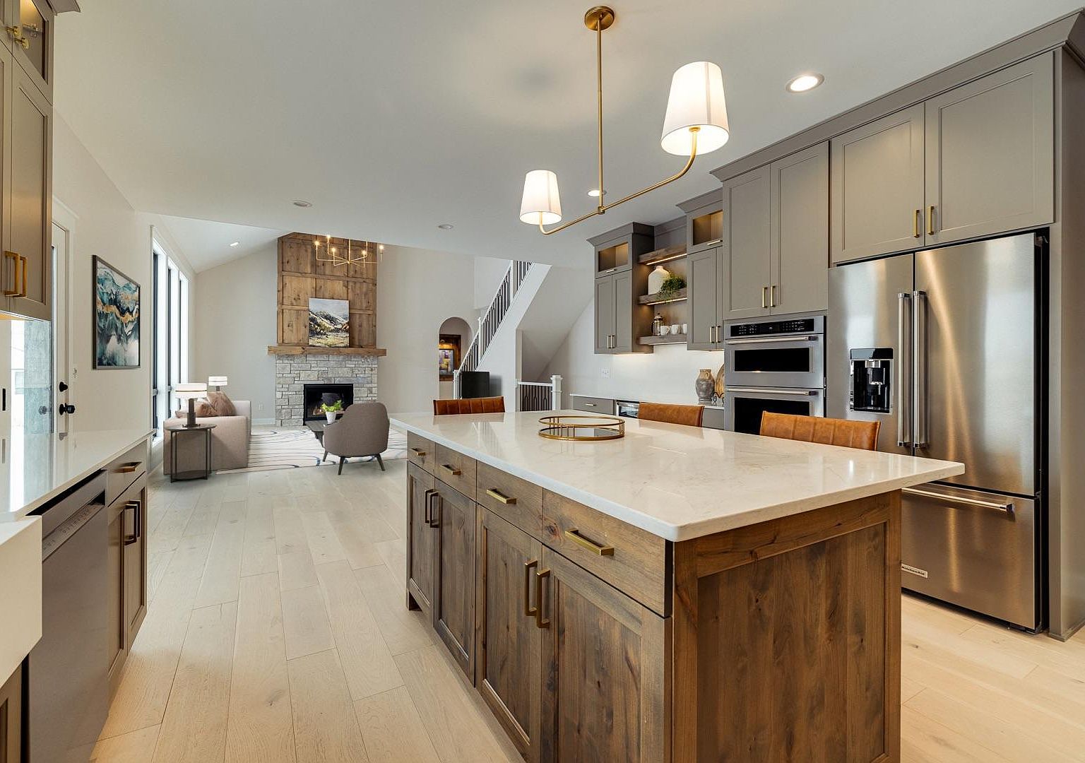 The gourmet kitchen at 2762 Mayo Prairie: an oversized stained-wood island with quartz counters, brass pendants, a stone surround range hood, double wall ovens, and warm-gray shaker cabinetry running back toward the great room